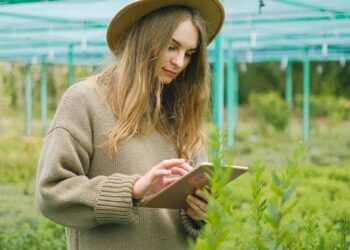 A young woman in a greenhouse using a tablet for plant care.
