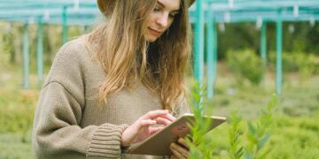 A young woman in a greenhouse using a tablet for plant care.