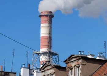 A tall industrial chimney emitting smoke, contrasting with urban rooftops under a bright blue sky.