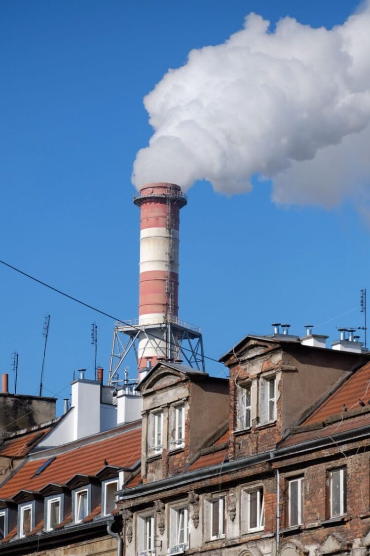 A tall industrial chimney emitting smoke, contrasting with urban rooftops under a bright blue sky.