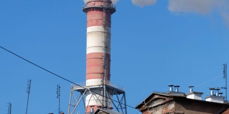 A tall industrial chimney emitting smoke, contrasting with urban rooftops under a bright blue sky.